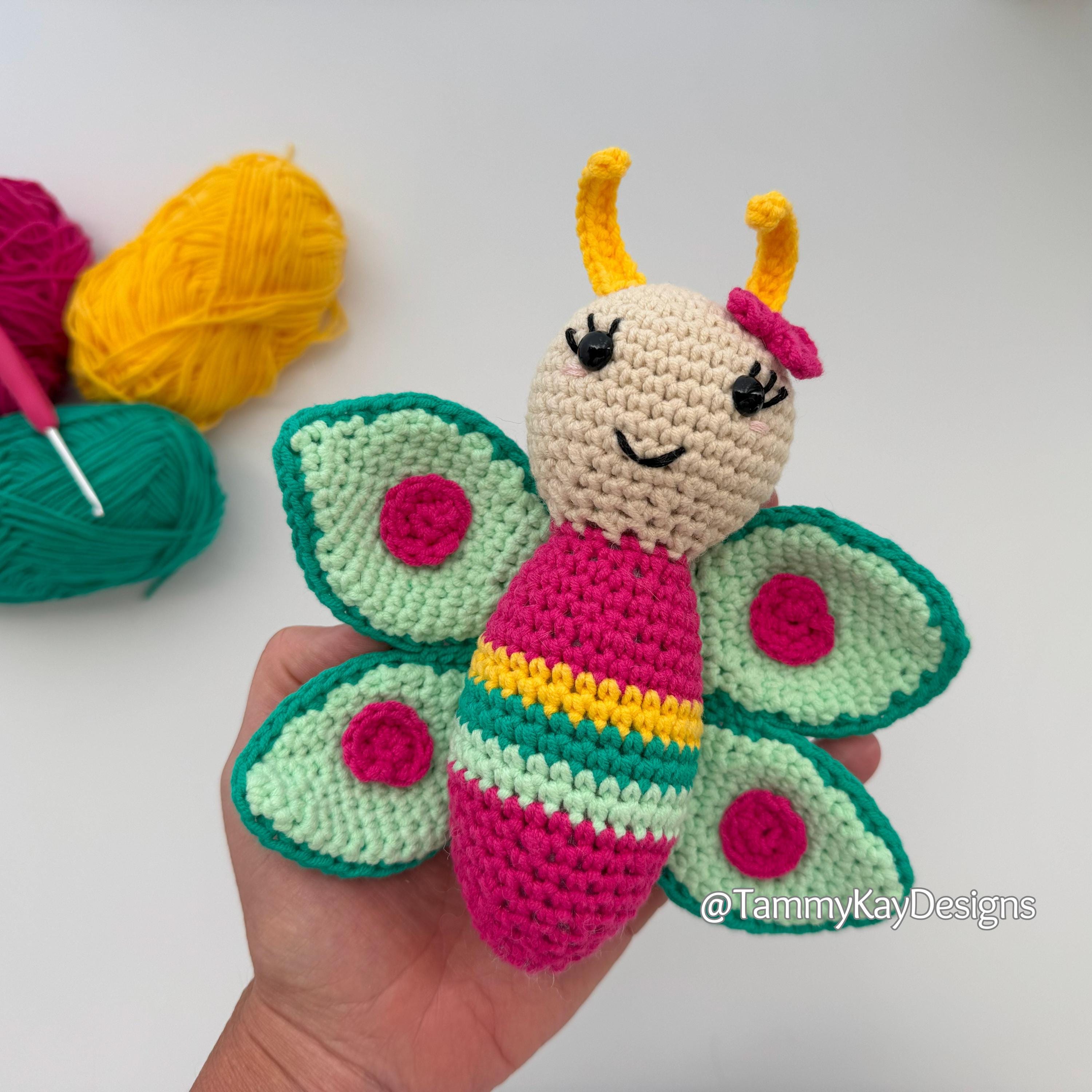 Hand holding a colorful crochet butterfly toy with yarn and a hook on a white background, featuring the brand 'Tammy Kay Designs'.
